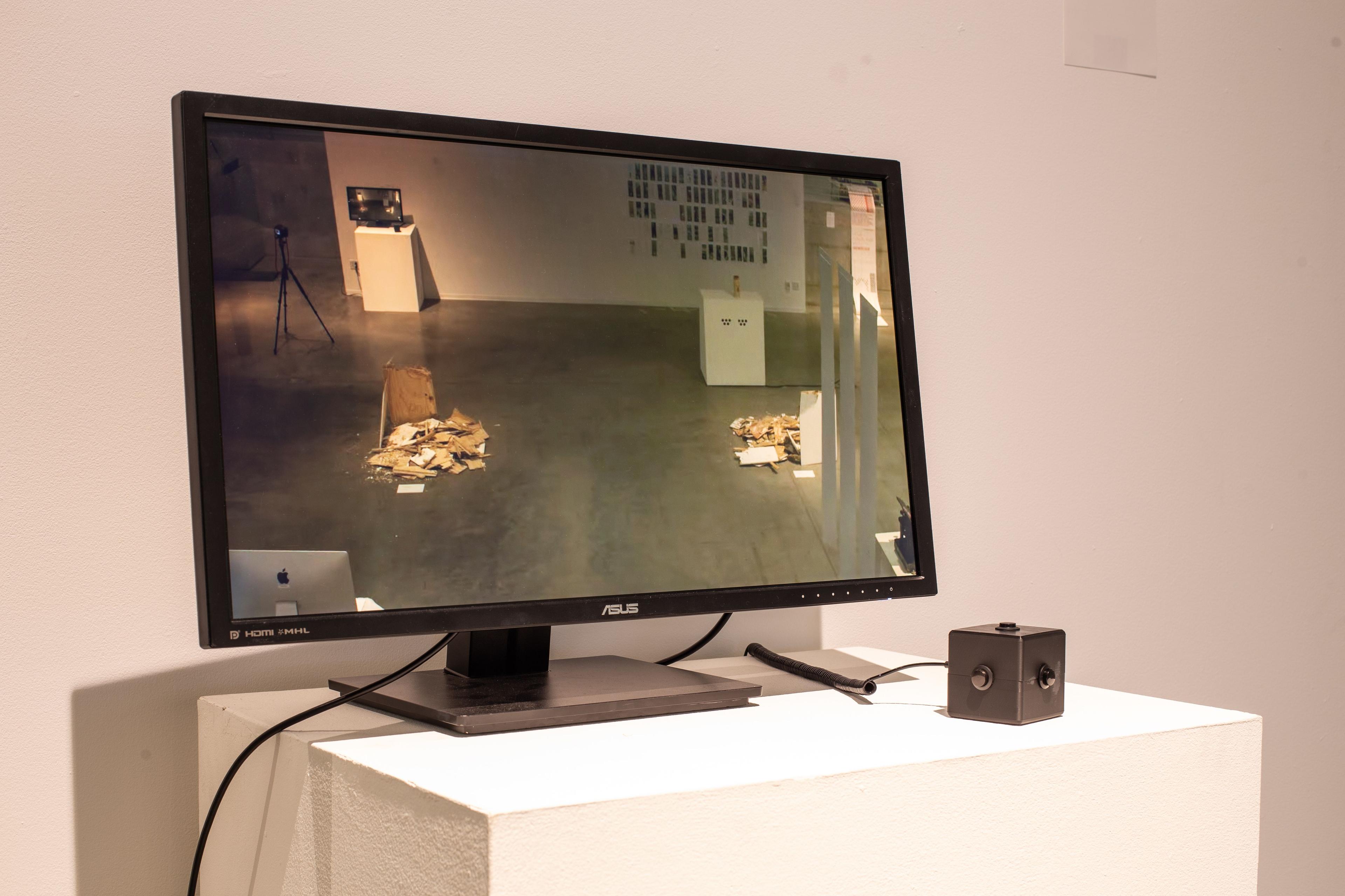 A monitor displays live footage of a camera positions on the ceiling behind the piece, showing viewers themselves within the exhibition space. Connected to the monitor is a cube-shaped controller, with a single button at the center of each face.