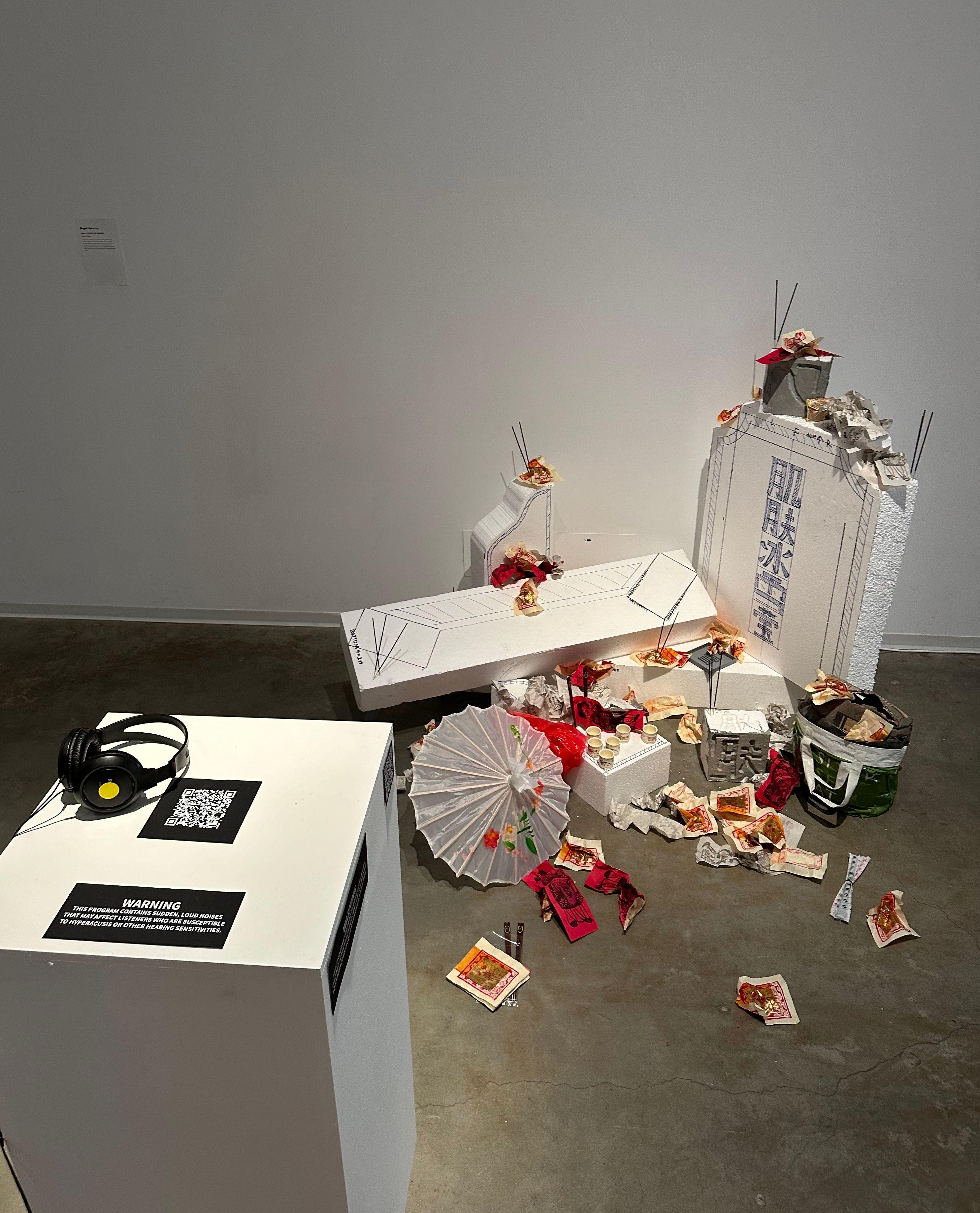 On the gallery floor styrofoam blocks with lines and characters drawn on them are arranged similar to a gravestone. Cups, incense sticks, paper napkins, and an umbrella cover the styrofoam blocks. A podium with a QR code, headphones, and a warning to part