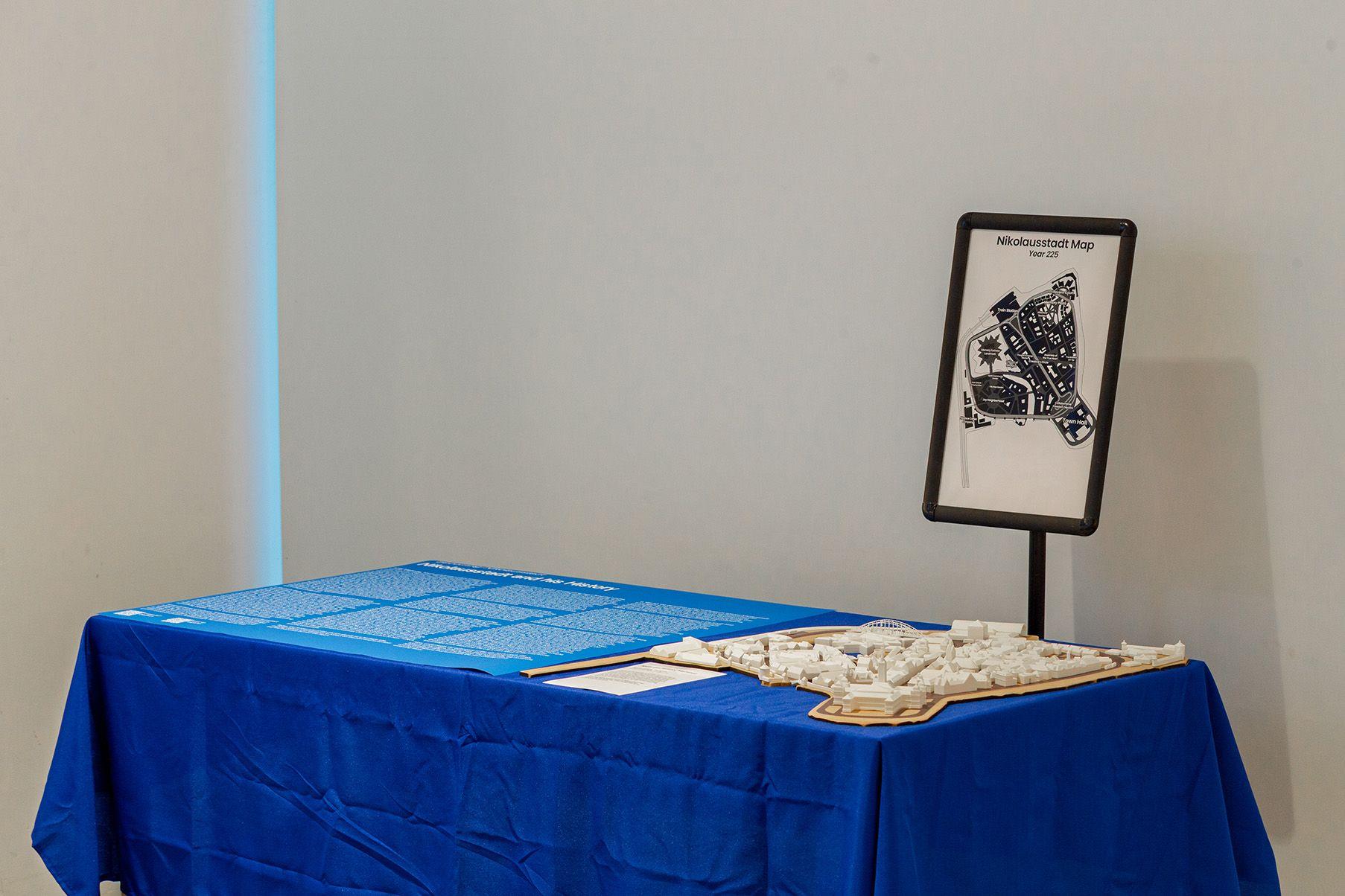 A tabletop exhibition display features a blue tablecloth, a large sheet with printed text, and a 3D model of a city layout mounted on a wooden base. To the right, a standing sign displays a black-and-white illustrated map titled "Nikolausstadt Map, Year 2