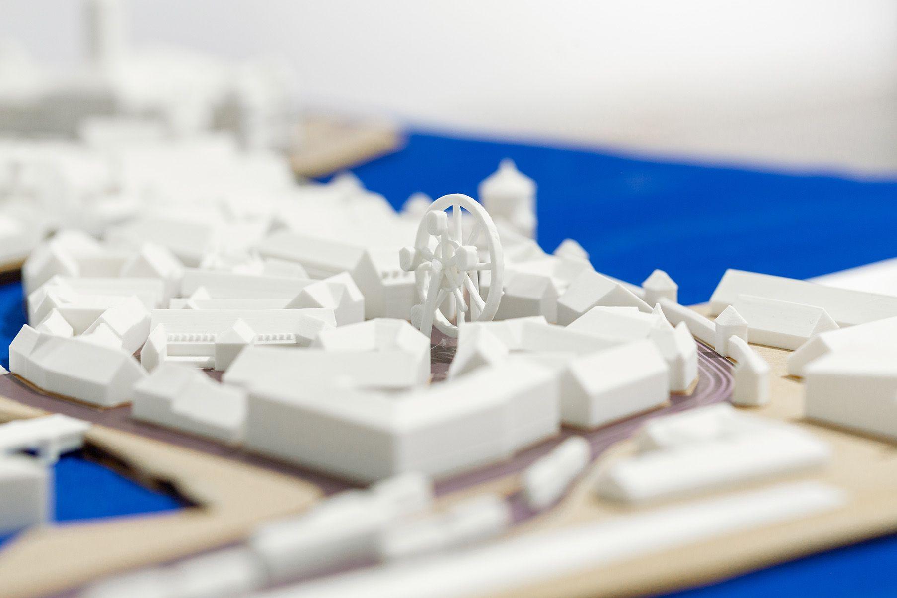 A close-up view of a white 3D-printed architectural model, showing a small urban area with clustered buildings. At the center stands the distinctive Ferris wheel-like structure. The model rests on a blue tablecloth, with roads and paths subtly indicated b