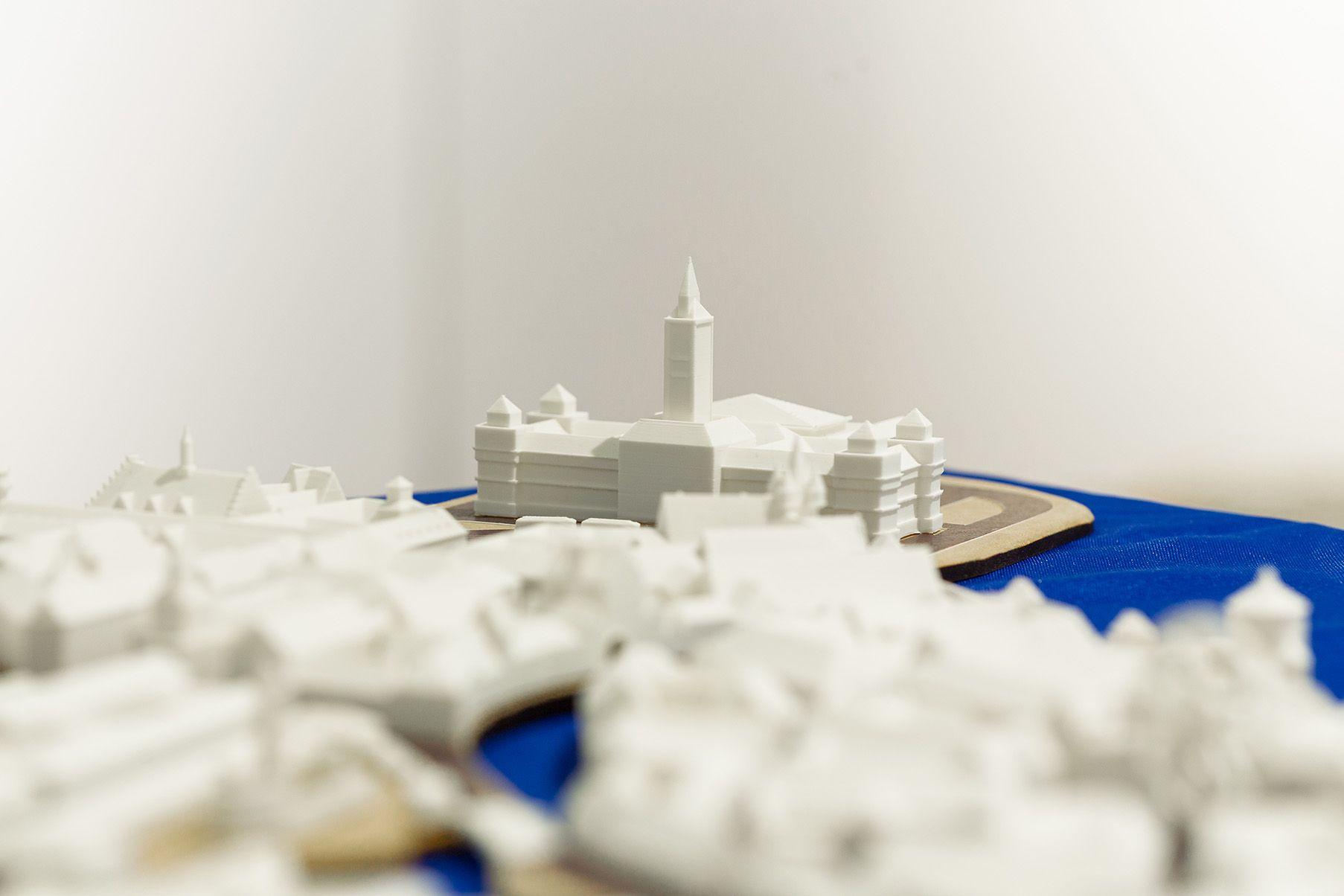 A central structure in a white 3D-printed model stands out with a tall tower and symmetrical wings, resembling the town hall. It is situated near a plaza. The surrounding area is filled with smaller model buildings, all rendered in white.