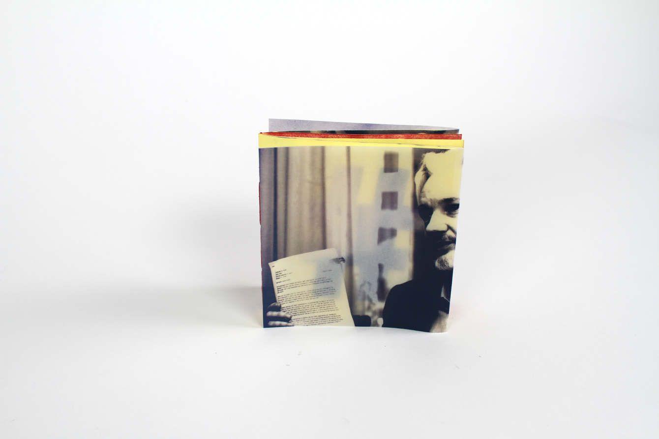 There is a square booklet, with only one side shown. On that page is an old man holding a document of text that is too small to read. The photo is in grayscale.