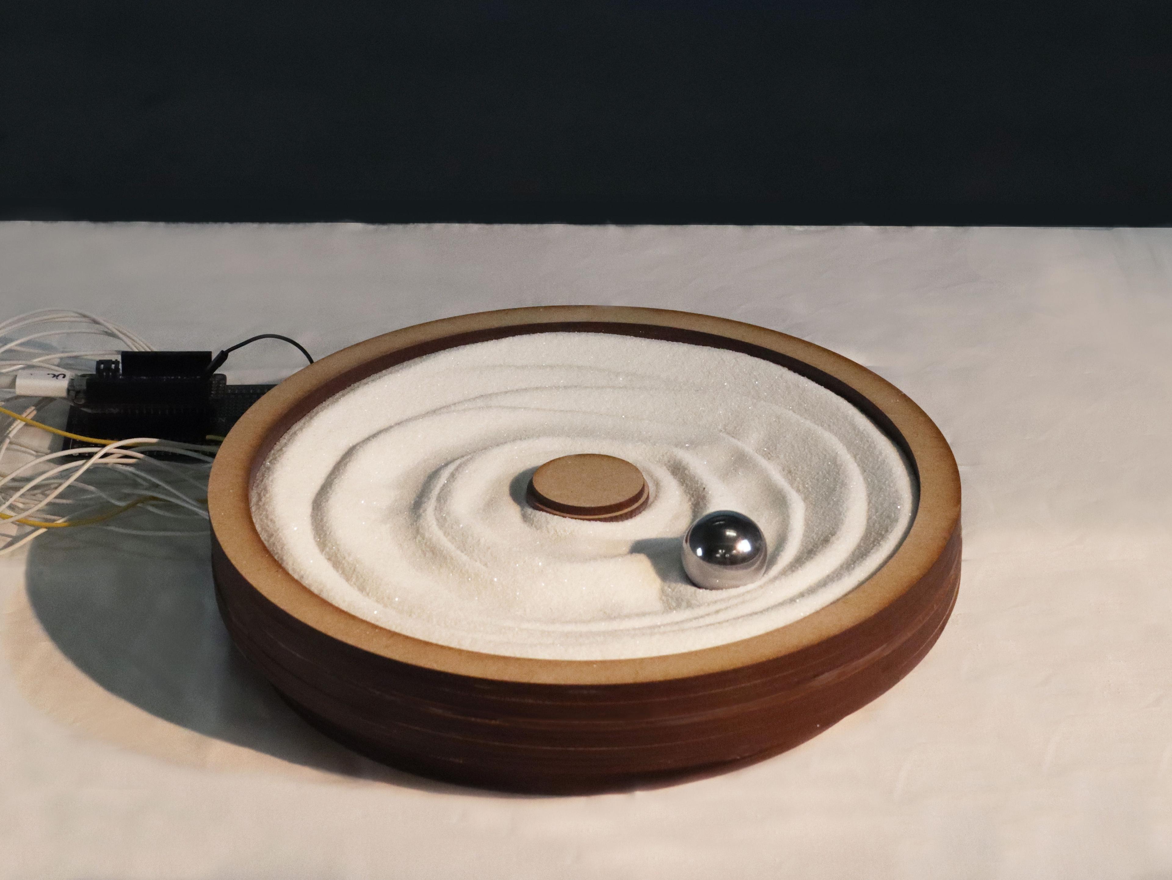 a round container of sand on a table. a metal ball is sitting in the sand. there are wires coming from the sand vessel.