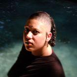 Person (Ren) looking at the camera over their shoulder. They are wearing a black shirt, and there is water in the background.