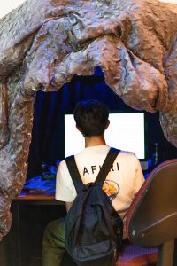 Catfishing video game with a sculpture housing it. Person is sitting inside the sculpture at a computer desk.