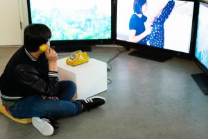 Person holding a yellow landline phone attached to triple monitor setup.