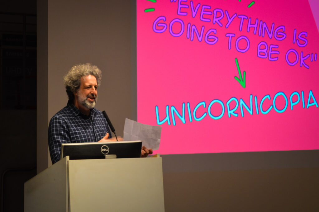 Edo Stern talks at a pedestal next to a slide that is projected displaying the word, Unicornicopia.