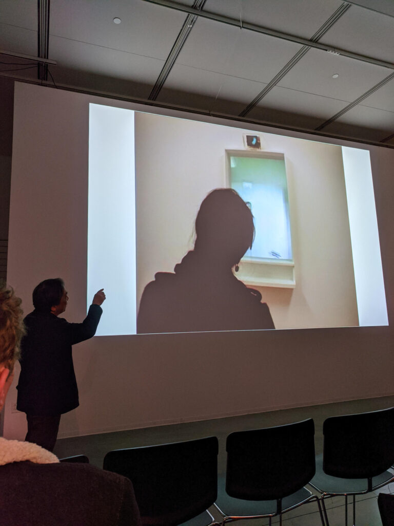 MASAKI FUJIHATA points to documentation of his work being projected on the walls of the EDA pit. The documentation is a person standing in front of a window.