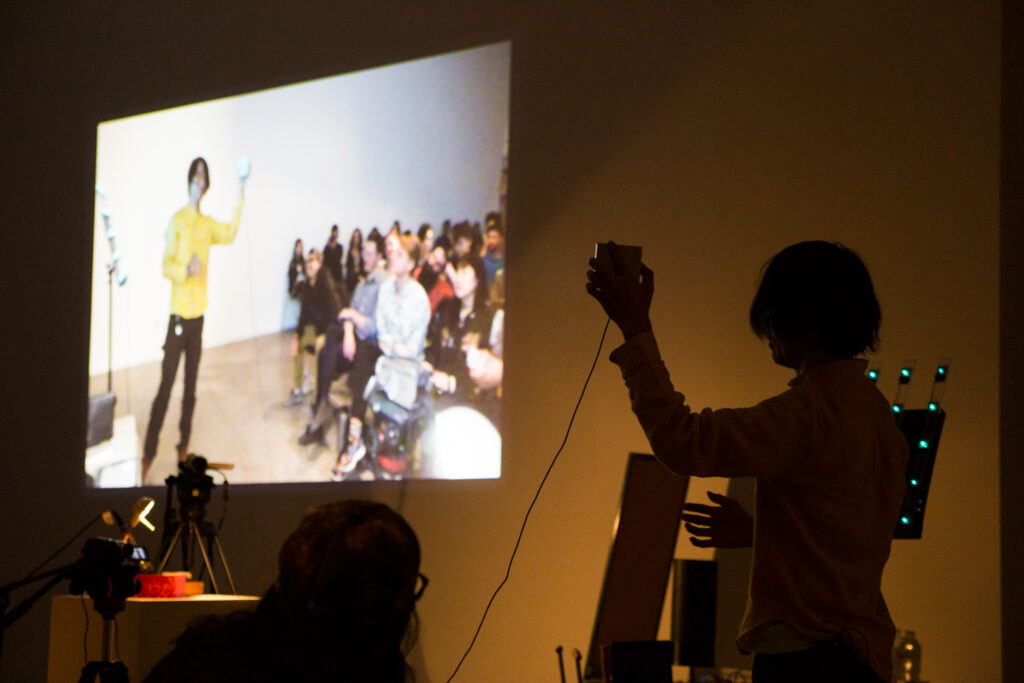 Toshio Iwai holding a camera that is capturing a live feed of the audience standing in front of Toshio.