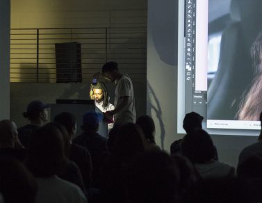 Yung Jake sitting at the podium as a friend illuminates him with his phone's flashlight.