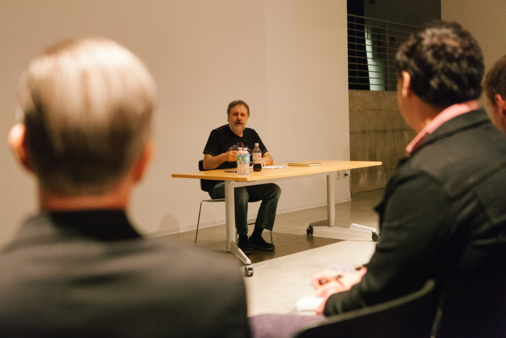 Zizek is sitting at a desk talking to an audience.