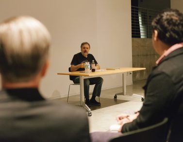 Slavoj Zizek talking to the crowd while sitting at a table.