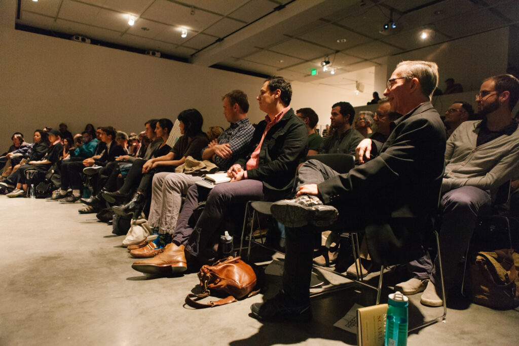 Audience members are sitting in front of Zizek. One of the audience members is sitting cross legged.