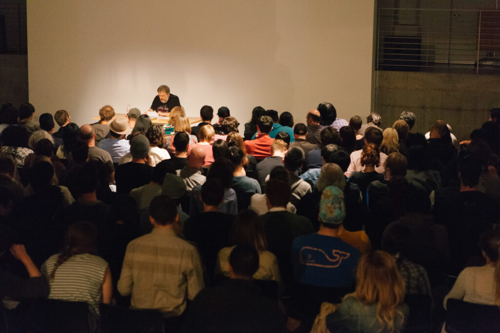 Zizek reading papers at a desk in front of an audience in the EDA pit.