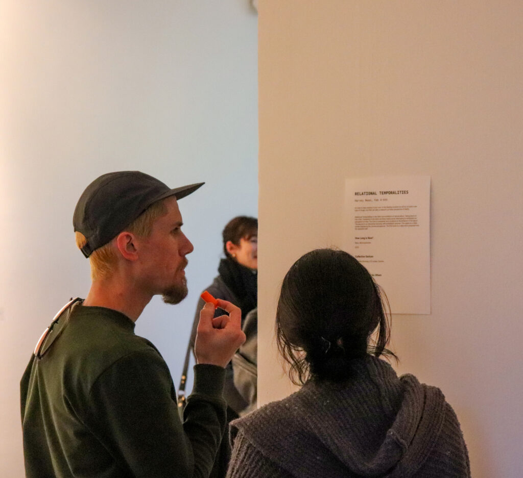 Gallery visitors look at the press release of Harvey Moon's Relational Temporalities mounted on the wall of the gallery.