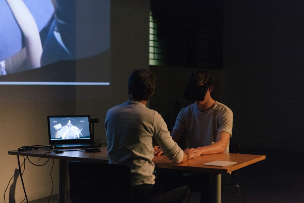 Person is wearing a VR headset and holding their hands out to another person on a desk.