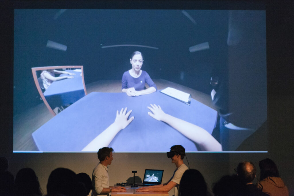 Person is wearing a VR headset and holding their hands out to another person on a desk.