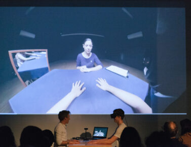 Person is wearing a VR headset and holding their hands out to another person on a desk.