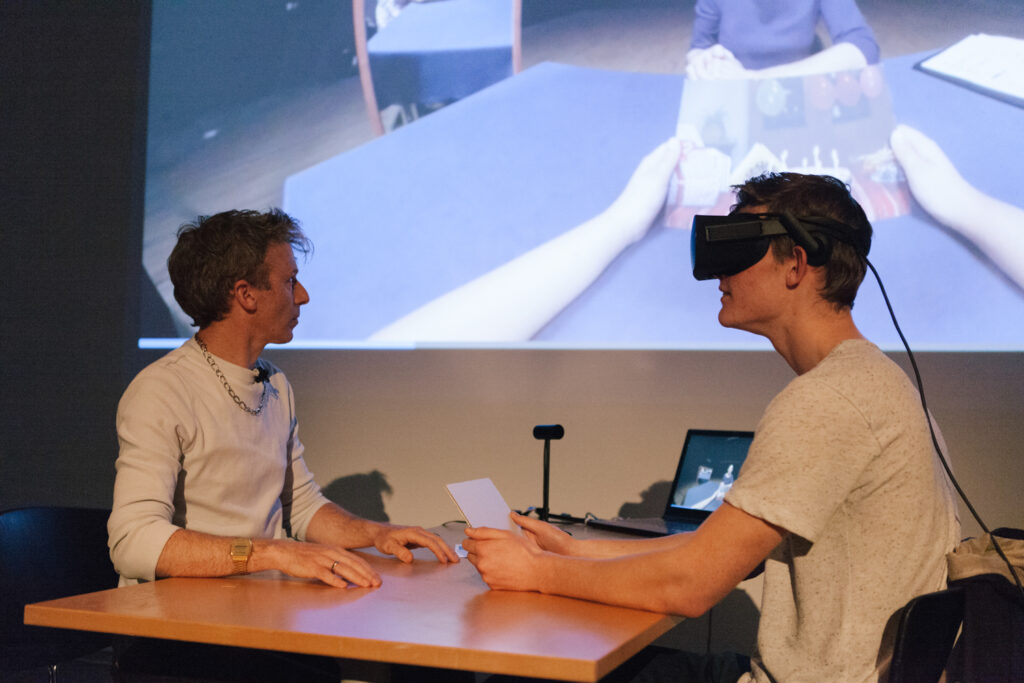 Person is wearing a VR headset and holding their hands out to another person on a desk.