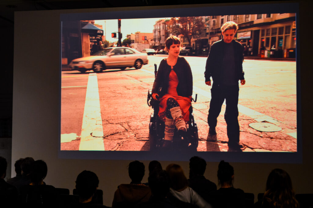 Movie still of a person in a wheel chair accompanied by a person walking beside the wheelchair person on the right.