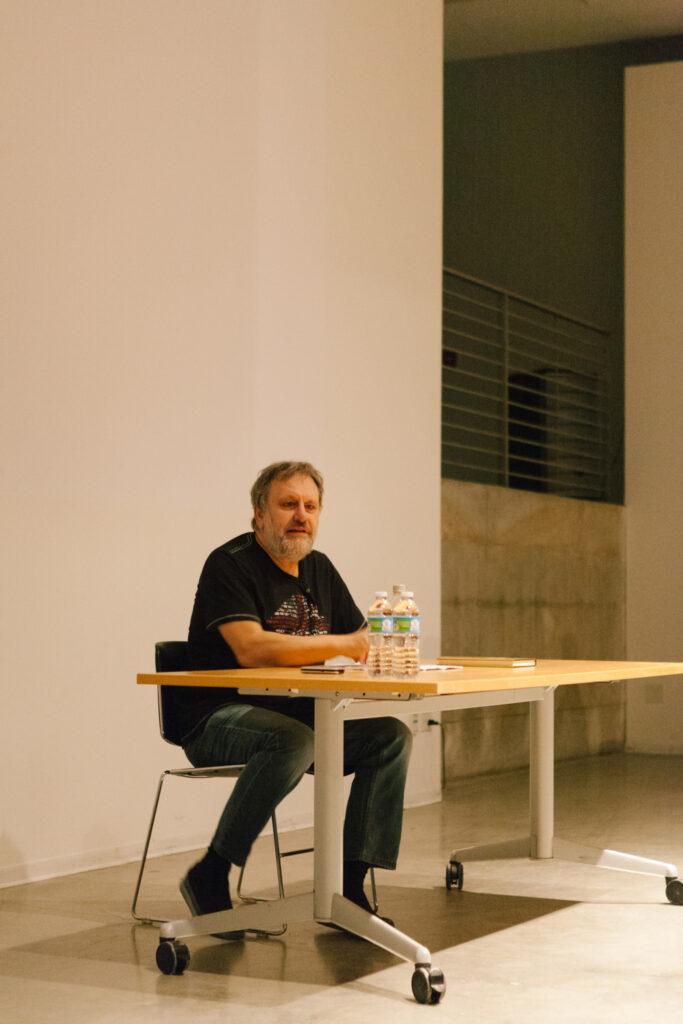 Zizek is sitting at a desk talking to an audience.