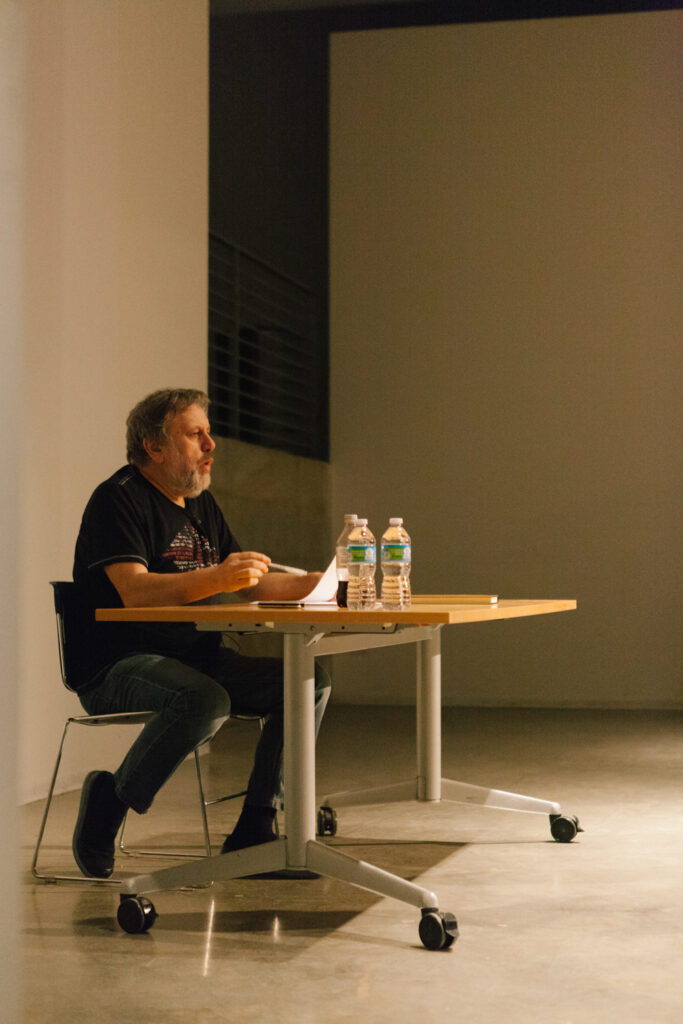 Zizek is sitting at a desk holding a pen and paper.